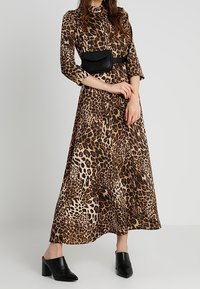 Abito maxi a stampa leopardata con maniche lunghe, collo alto e tessuto fluido. Indossato con una borsa a cintura nera e mules nere con tacco.