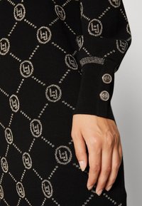 Hand som vilar på svart tyg med silver geometriskt mönster, ribbat manschett med två silverknappar som har cirkulära embleman.