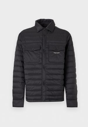 Homme debout, les mains dans les poches, portant une veste matelassée noire, un pull rouge, un pantalon gris, des chaussettes blanches et des mocassins marron foncé sur fond blanc.