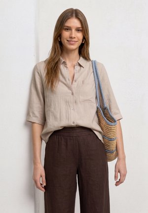 Jeune femme portant une chemise beige boutonnée et un pantalon marron foncé, tenant un sac en paille tissé avec des bretelles bleues sur l'épaule.