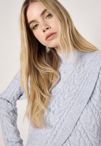 Jasnoniebieski sweter w warkocze z żebrowanym dekoltem i detalem po bokach. Miękka faktura z widocznym wzorem dzianiny i długimi rękawami.