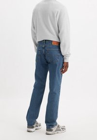 Jeans de mezclilla azul para hombre con un corte recto clásico, que cuentan con un parche de cuero de la marca, costuras visibles y una ligera textura desgastada.