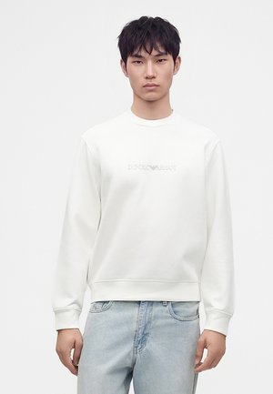 Νέος άνδρας με λευκό φούτερ Emporio Armani και ανοιχτόχρωμα τζιν, στέκεται μπροστά σε απλό λευκό φόντο.