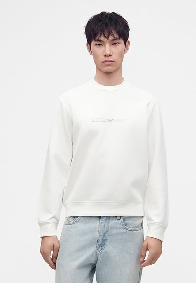 Νέος άνδρας με λευκό φούτερ Emporio Armani και ανοιχτόχρωμα τζιν, στέκεται μπροστά σε απλό λευκό φόντο.