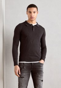 Mørkegrå langærmet polo-shirt med krave, fem-knaps placket og ribbede nederste kanter. Båret over en hvid T-shirt, parret med slidte jeans.