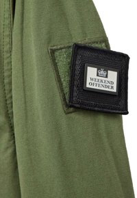 Olivenfarvet stof med en glat tekstur, der har en sort rektangulær lap med et hvidt "WEEKEND OFFENDER" logo og kroneemblemet.