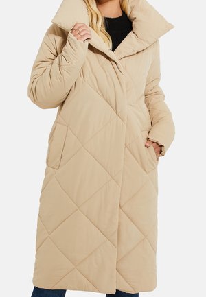 Femme portant un long manteau matelassé beige avec un grand col, des poches latérales et un top noir en dessous, posant avec une main dans la poche.