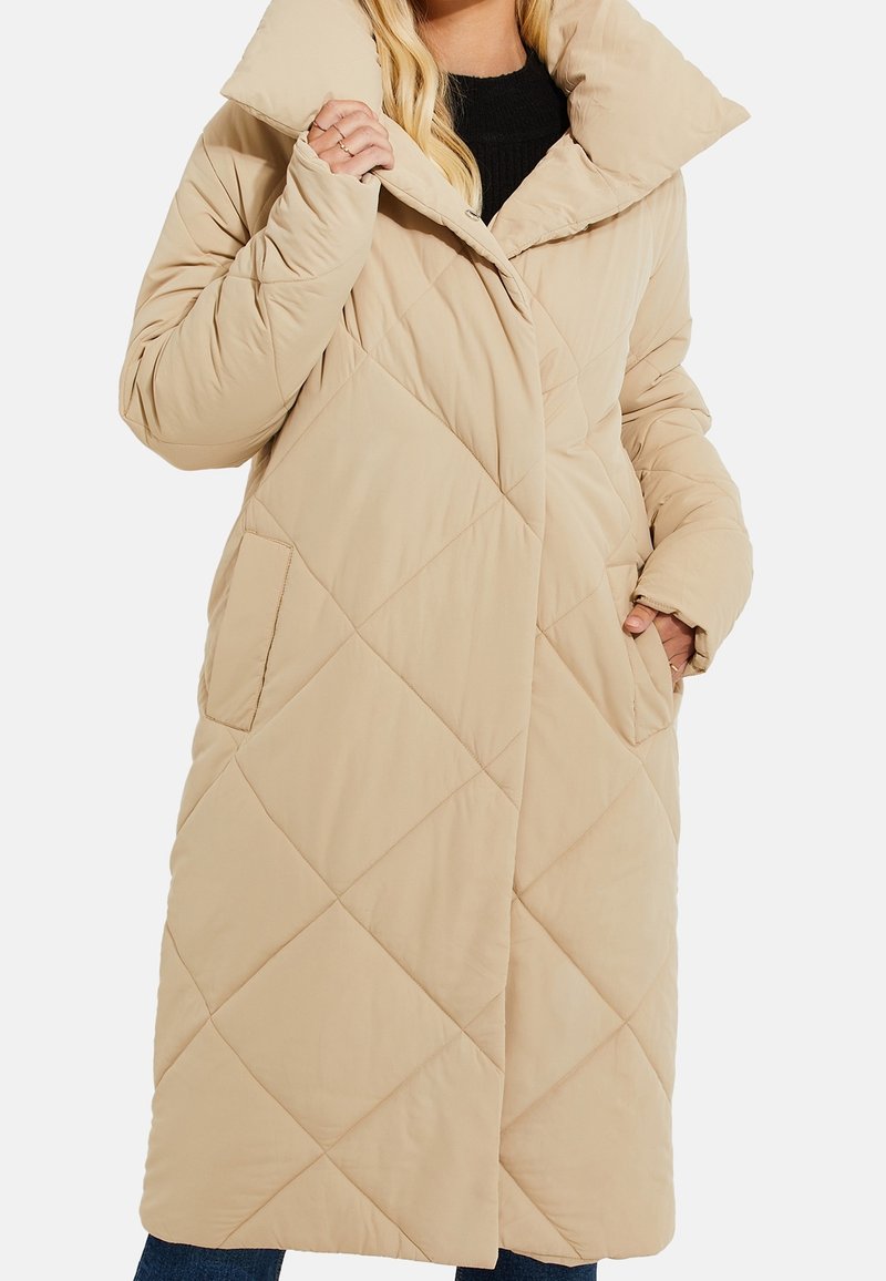Femme portant un long manteau matelassé beige avec un grand col, des poches latérales et un top noir en dessous, posant avec une main dans la poche.