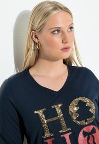 Marinblå V-ringad långärmad tröja med glittrande bokstäver i guld och rött. Modellen bär stora guldfärgade ringörhängen, mot en mjuk bakgrund.