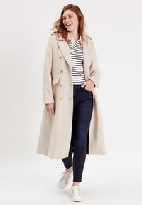 Manteau long beige avec des boutons dorés, des revers en pointe et des poches latérales, superposé sur une chemise rayée et un jean foncé, associé à des baskets blanches.