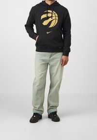 Sudadera negra con un gran gráfico de garra de baloncesto dorada, bolsillo canguro y capucha con cordón. Combinada con unos vaqueros claros desgastados y zapatos negros.