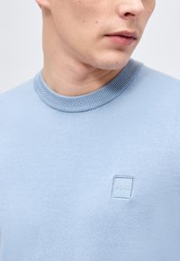 Jeune homme portant un pull bleu clair à col rond avec un logo carré en relief discret "BOSS" sur la poitrine, regardant sur le côté.