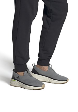 Personne portant des chaussures de sport grises à enfiler avec des semelles blanches et des accents noirs, associées à un pantalon jogger noir.