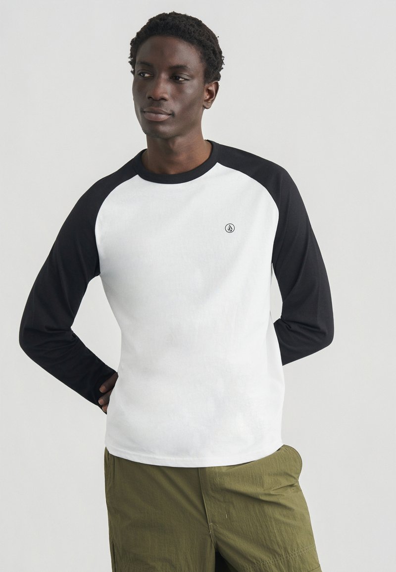 Μπλούζα με μανίκια ραγκλάν σε μαύρο χρώμα και λευκό σώμα, κατασκευασμένη από μαλακό βαμβάκι. Διαθέτει μικρό λογότυπο στο στήθος. Απλός, καθημερινός σχεδιασμός.