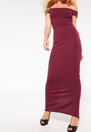 Femme portant une robe maxi ajustée bordeaux, épaules dénudées, avec des sandales à talons noires, debout devant un fond blanc uniforme.