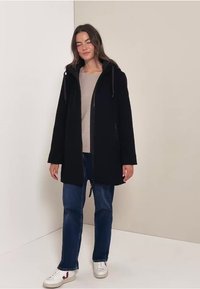Zwarte hooded jas met een rits, met een relaxte pasvorm, lange mouwen en zijzakken, gedragen over een lichte trui en jeans.
