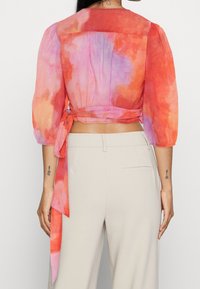 Blusa translúcida en tonos de naranja y rosa con mangas abullonadas de tres cuartos y detalle atado en la parte posterior, hecha de una tela liviana.