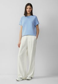 Lichtblauwe, kortlopende blouse met een ronde halslijn, gecombineerd met roomkleurige, wijde broek en grijze sneakers. Zachte stoftexturen zichtbaar.