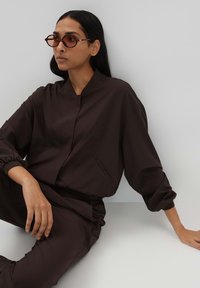 Brown, oversized top with a stand collar and elastic cuffs; matching pants with pockets; model wearing dark sunglasses, seated against a light wall.