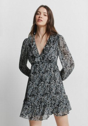 Młoda kobieta o długich brązowych włosach, ubrana w czarno-białą sukienkę z wzorem paisley, z długimi rękawami i falbaną na dole, stojąca na tle jednolitego tła.