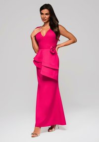 Nainen fuksia värisessä ilman hihoja oleva mekko, jossa on kerroksellinen peplum-yksityiskohta ja ruusukoriste, poseeraa yksi käsi vyötäröllä ja kultaisissa korkokengissä.