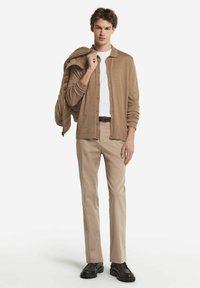 Beige strukturierte Strickjacke über einem weißen T-Shirt, kombiniert mit hellbeigen Hosen und dunklen Schuhen. Das Model posiert mit einer Jacke über einer Schulter.