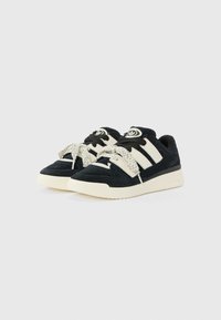 Black suede sneakers featuring white striped accents, textured laces, and a rubber sole with branding at the heel.