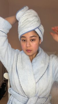 Person iført en blå badekåbe og et matchende håndklæde viklet omkring hovedet, justerer håndklædet med den ene hånd indendørs.