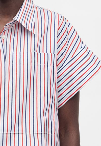 Πουκάμισο με κοντό μανίκι με κόκκινες, μπλε και λευκές κάθετες ρίγες και τσέπη στο στήθος σε σκουρόχρωμο χέρι σε ουδέτερο φόντο.
