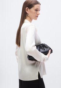 White satin blouse with a collar, long sleeves, and side slits, paired with a black gathered clutch. Smooth texture and elegant design.