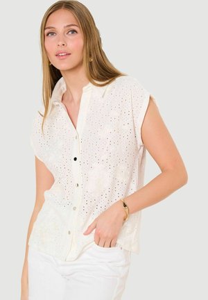 Noor naine, kes kannab varrukateta valget augulist pluusi ja valgeid pükse, pikkade sirgete juuste ja kuldse käevõruga, poseerimas lihtsa tausta ees.