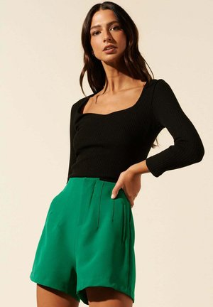 Haut noir à manches longues côtelées avec un décolleté carré, associé à un short plissé vert. Le short a une texture lisse et une coupe décontractée.
