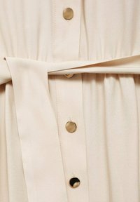 Abito beige con un davanti a bottoni, caratterizzato da bottoni in tonalità oro, una cintura annodata e una texture plissettata lungo la vita per un dettaglio di design aggiuntivo.