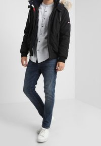 Muž v černé kapuci s kožešinovým lemem, světle šedé košili na knoflíky, modrých džínách a bílých teniskách stojící v neutrálním prostoru.