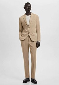 Beigefarbener Anzug mit einem Zweiknopf-Blazer, abgesteppten Revers und schmal geschnittenen Hosen. Komplettiert mit einem weißen Rundhalshemd und schwarzen Schuhen.