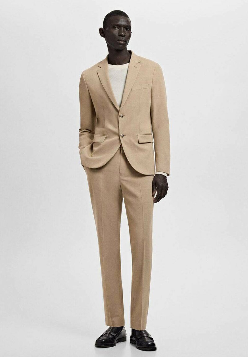 Beige suit with a two-button blazer, notched lapels, and slim-fit trousers. Complemented by a white crew-neck shirt and black shoes.