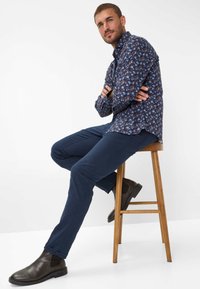 Hemd im Marine-Muster, dunkelblaue eng anliegende Hose und braune Knöchelh Boots. Das Hemd hat florale Designs und einen lockeren Schnitt.