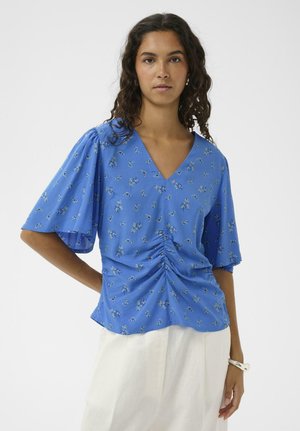 Žena s tmavými kučeravými vlasmi, oblečená v modrej kvetinovej blúzke s výstrihom do V a zberaným predným dielom, a bielych nohaviciach, stojaca pred jednoduchým svetlým pozadím.