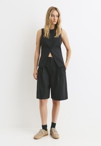 Dunkel gestreifter ärmelloser Weste mit hohem Kragen und fünf Knöpfen, kombiniert mit locker sitzenden dunklen Shorts. Beigefarbene Sneakers vervollständigen den Look.