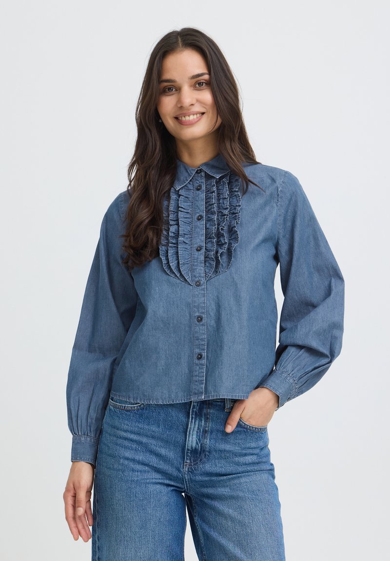 Jeune femme souriante, portant une chemise en jean bleue avec des volants sur le devant et un jean bleu, posant avec une main dans la poche.
