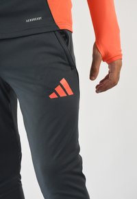 Pantalones deportivos grises con una textura suave y un prominente logo rojo de Adidas en el muslo, combinados con una camiseta de manga larga naranja.