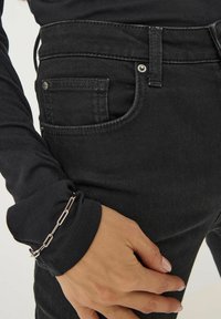 Czarne dżinsy z wysokim stanem mają subtelne nity, tonalne przeszycia oraz klasyczny pięciokieszeniowy design. Widoczna jest również czarna koszula z długim rękawem oraz srebrna bransoletka.