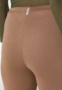 Hnědé legíny vyrobené z elastického materiálu, se šitým designem, bezšvovými boky a malou nášivkou umístěnou poblíž pasu.