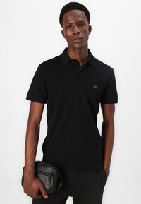 Polo preto com colarinho clássico e mangas curtas. Apresenta um logótipo discreto no peito. Acompanhado de uma bolsa preta com fecho éclair.