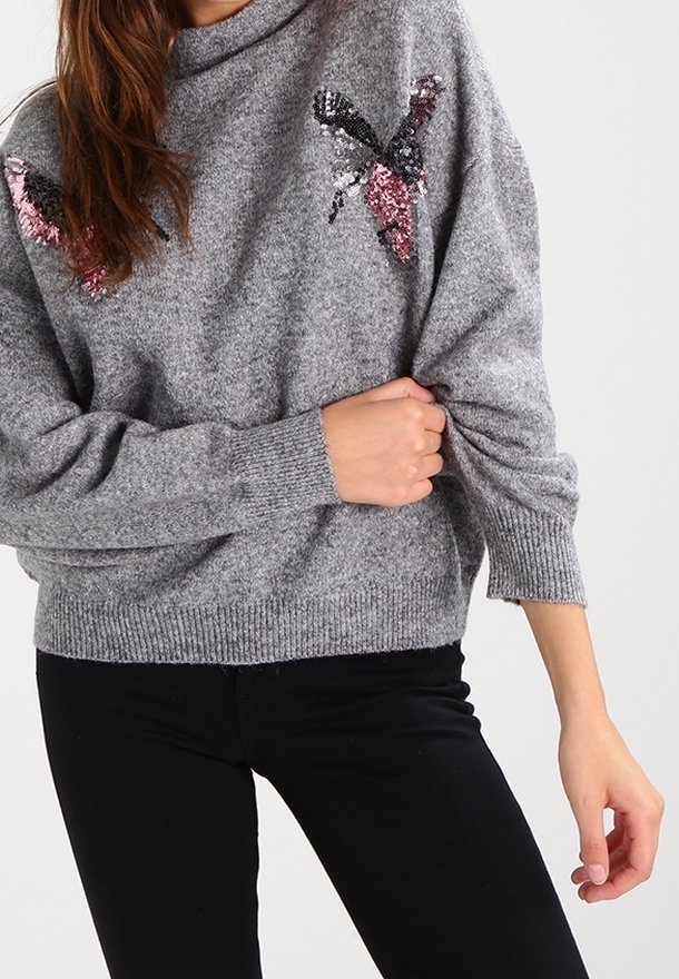 Femme portant un pull gris confortable avec des dessins de papillons en sequins sur les épaules, tenant légèrement une manche, associé à un pantalon noir.
