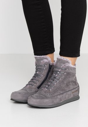 Bottes montantes en daim gris avec doublure en fourrure, lacets et fermeture éclair latérale, portées avec un pantalon noir ajusté sur un fond uni.