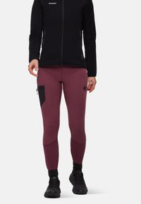 Leggings bordeaux con un design aderente, dotati di una tasca con zip nera, abbinati a una giacca nera con zip e calzature nere.
