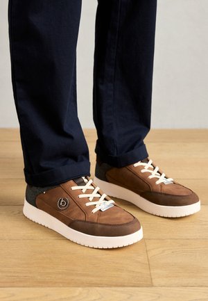 Hnedé tenisky vyrobené z texturovanej látky s krémovými šnúrkami, bielymi podrážkami a šedým akcentom na päte. Detail loga na boku.