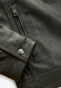 Close-up of dark leather jacket sleeve with a metal snap button labeled "Signature Goods Durable" and a vertical zippered pocket.