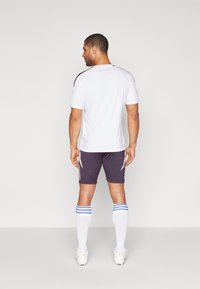 Jersey atlético blanco con acentos negros, combinado con pantalones cortos oscuros que tienen rayas blancas. Lleva calcetas blancas hasta las rodillas y botas de fútbol.